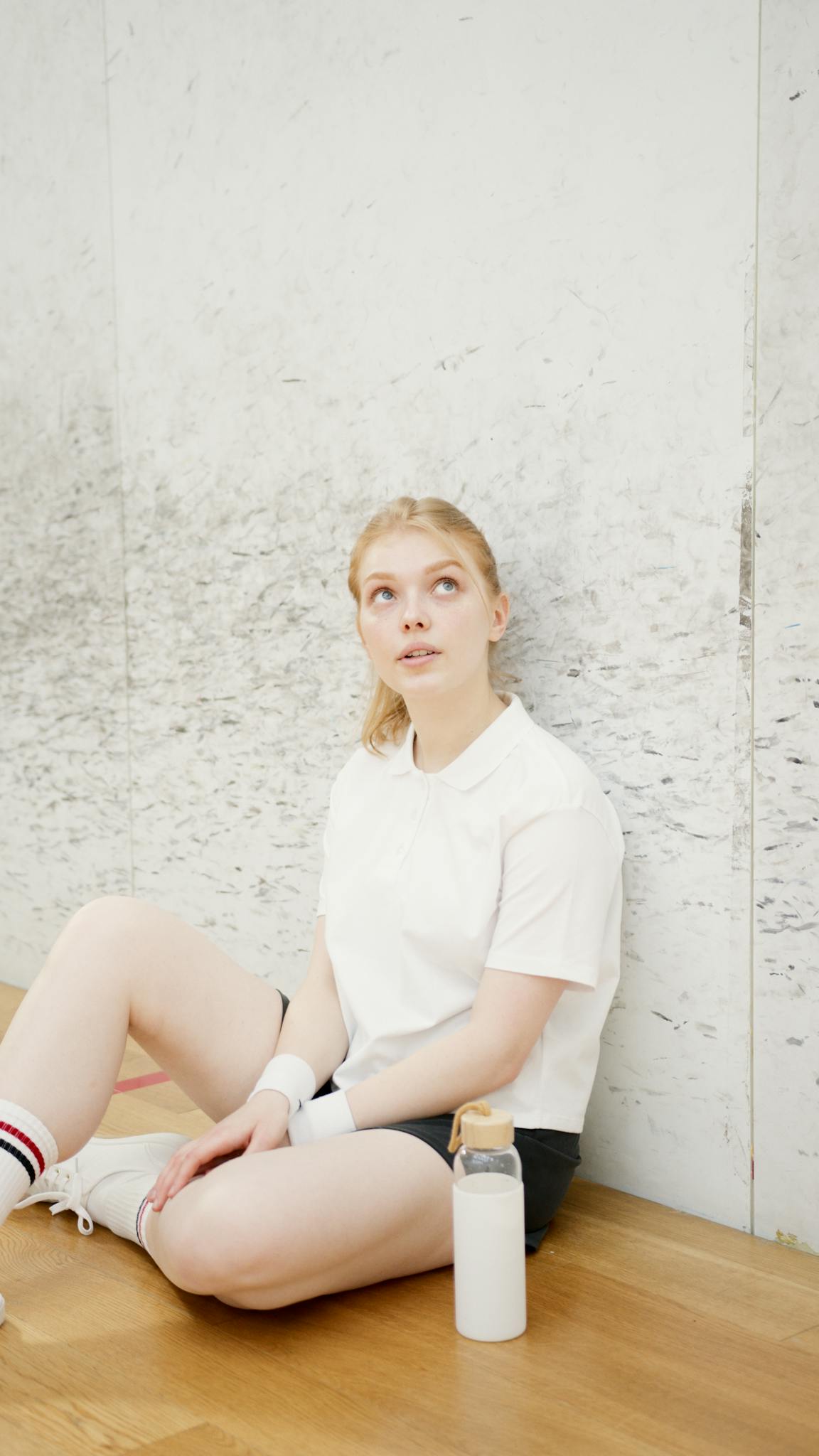 Caucasian woman athlete resting indoors on wooden floor in sports attire with water bottle.